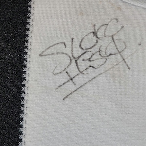 May include: Close-up of a white fabric with a black border. The fabric has a handwritten signature in dark gray ink. The signature appears to read "Sleep". The fabric has a subtle texture.