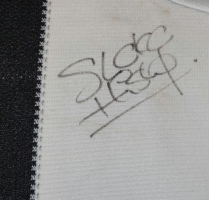 May include: Close-up of a white fabric with a black border. The fabric has a handwritten signature in dark gray ink. The signature appears to read "Sleep". The fabric has a subtle texture.