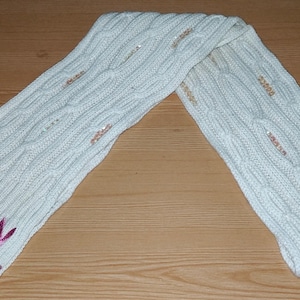 Op de afbeelding: Crèmekleurige kabelgebreide sjaal met roze rand en geborduurd konijnenlogo. De sjaal heeft een textuurpatroon en is versierd met kleine pailletten. Een stijlvol winteraccessoire voor warmte.