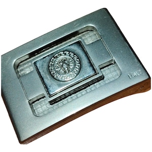 K&ouml;nnte beinhalten: Silberfarbene Metallschnalle mit rechteckigem Design. Die Schnalle hat ein zentrales Quadrat mit einem erhabenen kreisf&ouml;rmigen Emblem. Das Emblem zeigt ein detailliertes Relief eines Gesichts, umgeben von einem dekorativen Rand. Das Wort "ITALY" ist sichtbar.