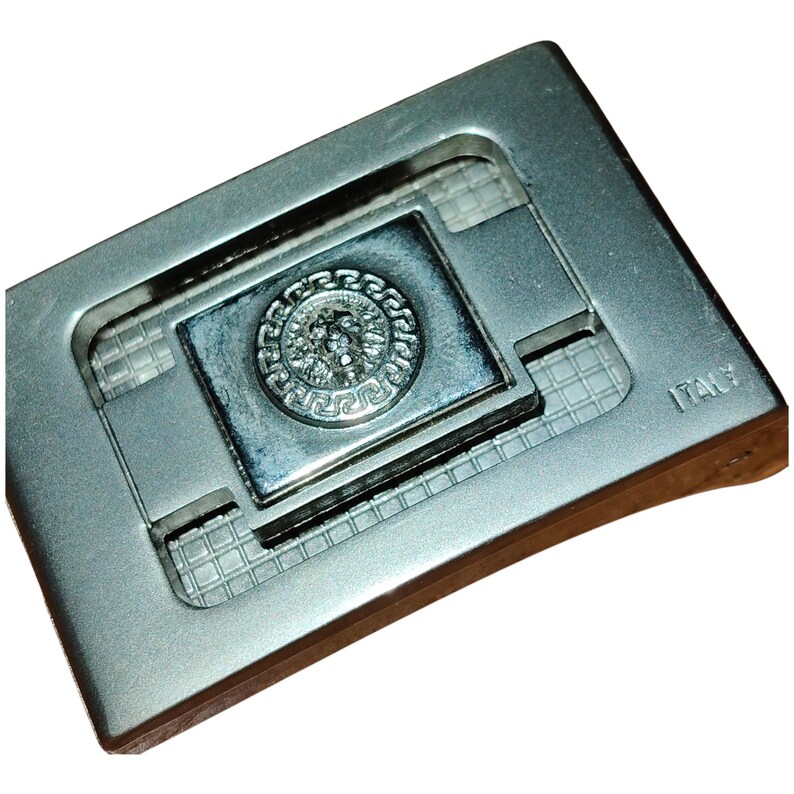 K&ouml;nnte beinhalten: Silberfarbene Metallschnalle mit rechteckigem Design. Die Schnalle hat ein zentrales Quadrat mit einem erhabenen kreisf&ouml;rmigen Emblem. Das Emblem zeigt ein detailliertes Relief eines Gesichts, umgeben von einem dekorativen Rand. Das Wort "ITALY" ist sichtbar.