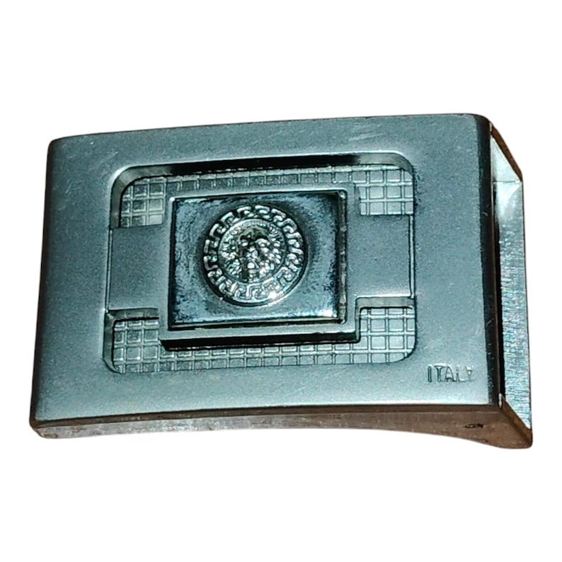 K&ouml;nnte beinhalten: Silberfarbene Metallschnalle mit rechteckigem Design. Die Schnalle hat ein zentrales Emblem mit kreisf&ouml;rmigem Muster und detailliertem Design. Das Wort "ITALY" ist auf der rechten Seite der Schnalle zu sehen.