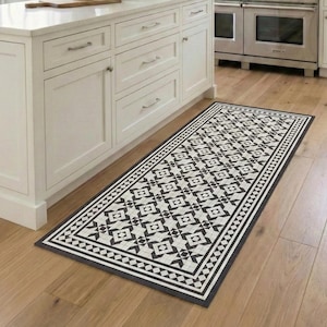 Może przedstawiać: Prostokątny dywan kuchenny w czarno-biały wzór z geometrycznym designem. Dywan leży na podłodze z twardego drewna w jasnej, białej kuchni. Wymiary dywanu to około 60 cm szerokości i 183 cm długości.