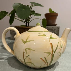 May include: Cream ceramic teapot with a rounded body, decorated with a bamboo design in green and brown. It has a curved handle, a spout, and a lid with a green finial. A decorative item for the kitchen.