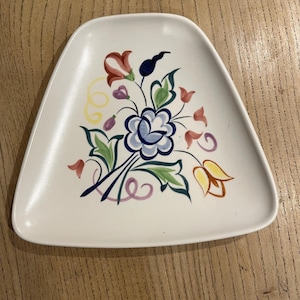 Puede incluir: Un plato blanco triangular con un diseño floral pintado a mano. Las flores son en tonos de azul, rojo, amarillo y morado, con hojas y tallos verdes. El plato está sobre una superficie de madera.