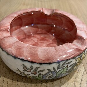 Könnte beinhalten: Ein runder Aschenbecher aus Keramik mit einem rosa-weißen, craquelé glasierten Rand und einem floralen Muster am Boden. Der Innenraum ist tiefrot-rosa. Der Aschenbecher steht auf einer Holzoberfläche.
