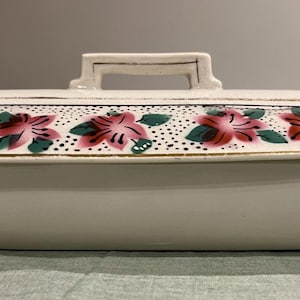 May include: A white ceramic serving dish with a lid. The lid is decorated with pink floral designs and green leaves. The dish has a rectangular shape with a handle on top. The dish is likely vintage.
