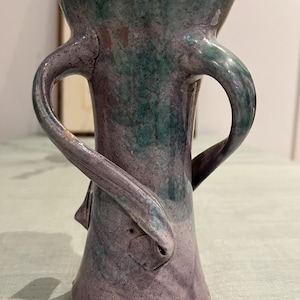 Puede incluir: Un jarrón de cerámica hecho a mano con un diseño único. El jarrón presenta un cuerpo cilíndrico con dos asas curvas y una parte superior ensanchada. La paleta de colores incluye tonos de morado, verde y marrón, creando una apariencia texturizada y moteada. El jarrón mide aproximadamente 20 cm de alto.