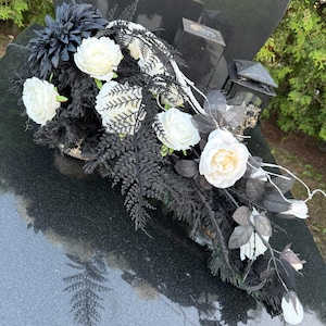 Könnte beinhalten: Ein Grabgesteck mit einer Mischung aus weißen Rosen und Dahlien, die mit schwarzen künstlichen Farnen und Blattwerk kontrastieren. Das Dekorationsobjekt ist kaskadenförmig gestaltet und enthält dunkle Stiele und Blätter.
