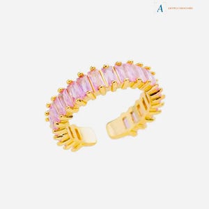 Puede incluir: Un anillo ajustable de color dorado con una fila de piedras rectangulares de color rosa claro. El anillo tiene una banda texturizada y está abierto en la parte inferior. El anillo está sobre un fondo blanco. El texto "ARTPLUSHSTORE" está en la esquina superior derecha.