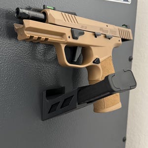 Könnte beinhalten: Hellbraune Pistole mit schwarzem Magazin, montiert auf einer schwarzen Halterung an einer grauen Oberfläche. Die Pistole hat einen strukturierten Griff und der Text "Sig Sauer P322" ist sichtbar. Ein weißes Etikett mit Text befindet sich im Hintergrund.