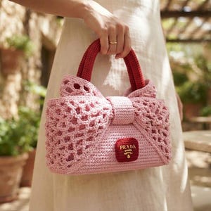 Puede incluir: Un bolso de mano de ganchillo rosa claro con un diseño de lazo estructurado, dos asas superiores de color rojo oscuro y un parche rectangular central de color rojo oscuro con el logotipo de Prada en letras doradas y un patrón de malla calada.