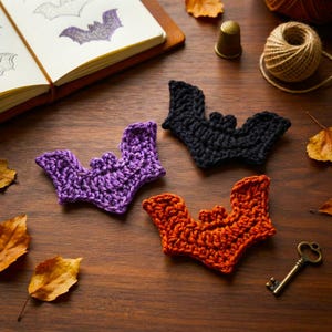 May include: Three crocheted bat appliques in purple, black, and orange yarn. Each bat features a distinct wing shape and small ears. The items are arranged on a wooden surface alongside a notebook with bat sketches, a thimble, twine, and a key.
