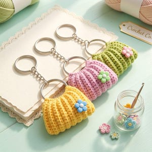 Könnte beinhalten: Drei gehäkelte Schlüsselanhänger in Form von Miniatur-Handtaschen in Gelb, Helllila und Hellgrün. Jeder verfügt über einen silbernen Metallring und eine kleine, einfarbige Blumenverzierung auf der Vorderseite des strukturierten Garnbeutels.