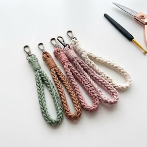 Op de afbeelding: Vier gevlochten macramé polsband-sleutelhangers in saliegroen, bruin, oudroze en crème. Elke sleutelhanger heeft een donker metalen karabijnhaak bevestigd aan een lus van geweven katoenen touw, gerangschikt in een rij naast een haaknaald.