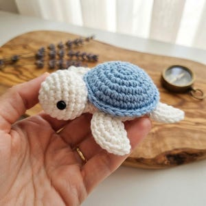 May include: A small, handmade crochet turtle toy featuring a white head, white flippers, and a textured light blue shell. The turtle has a single black bead eye visible on the side of its head, crafted with detailed yarn stitching.