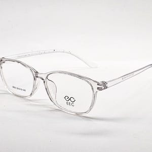 Puede incluir: Gafas rectangulares transparentes con montura transparente. Las gafas presentan un diseño clásico con puente y patillas visibles. Las lentes son transparentes y la montura tiene el texto "2022 52015-142" y "EEC" impreso. El estilo general es moderno y minimalista.