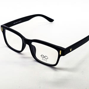 Puede incluir: Gafas negras con montura rectangular y lentes transparentes. Las gafas tienen un acabado mate y detalles dorados en las patillas. La marca "EEC" está impresa en la lente.
