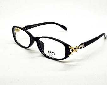 Könnte beinhalten: Schwarze Brille mit ovalen Gläsern und goldenen Akzenten an den Bügeln. Die Brille hat ein modernes Design, mit dem Markennamen "EEC" auf dem Glas.