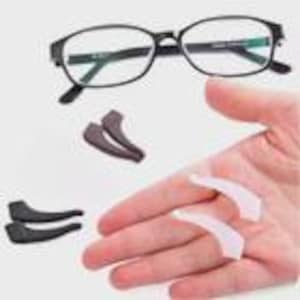 Puede incluir: Gafas con montura negra y lentes ovaladas. Se muestran dos pares de almohadillas de silicona para las orejas, de color marrón oscuro y blanco. Las almohadillas están diseñadas para evitar que las gafas se deslicen. Las gafas y las almohadillas están sobre un fondo claro.