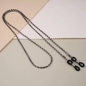 May include: A dark gray metal eyeglass chain with a textured, linked design. The chain has black rubber loops at each end to attach to glasses. The chain is shown against a neutral background.