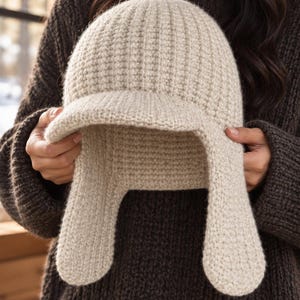 May include: A beige knit hat featuring a ribbed texture, a short brim, and two long ear flaps extending downward. The hat is constructed from a thick, soft yarn material with a consistent light tan color throughout the entire piece.
