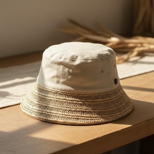 May include: A beige bucket hat with a woven straw brim. The hat is made of a light-coloured fabric and features a dark button detail. The hat is on a wooden surface, suggesting a casual, everyday accessory.