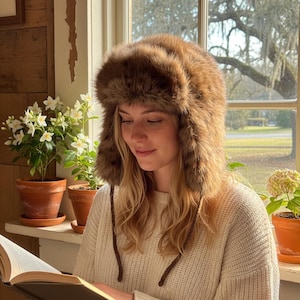 May include: A brown faux fur trapper hat with ear flaps. The hat is worn by a person reading a book indoors. The person is wearing a cream-colored sweater. Potted plants are on a windowsill near a window with a view of trees.