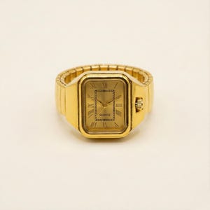 Puede incluir: Un reloj de anillo dorado con una esfera rectangular y números romanos. La esfera del reloj dice "QUARTZ". La correa es de estilo flexible y extensible. El reloj es de estilo vintage.