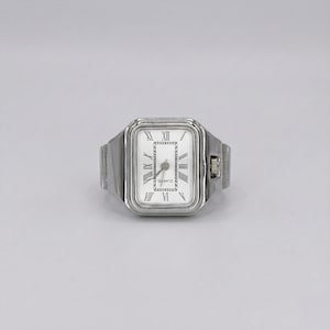 Può includere: Un orologio ad anello argentato con quadrante rettangolare bianco. Il quadrante presenta numeri romani e lancette nere, con la scritta "QUARTZ" stampata sotto. L'anello ha un design semplice e moderno.