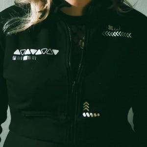 Könnte beinhalten: Schwarze Jacke mit Reißverschluss und weißen geometrischen Designs sowie dem Text "LA STRIA" unten. Die Jacke hat einen hohen Kragen und einen Frontreißverschluss. Das Design umfasst Dreiecke, Pfeile und andere Formen.