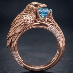 May include: A rose gold ring featuring a sculpted raven with detailed feathers. The bird holds a round, faceted light blue gemstone in its beak. The band is decorated with small, alternating clear and pink stones set along the sides.