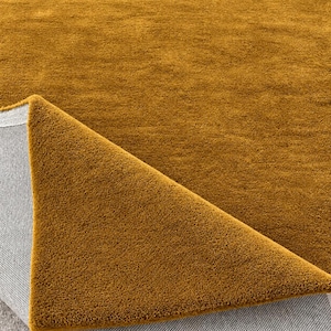 May include: A solid mustard yellow area rug with a dense, short-pile texture. One corner of the rug is folded over to reveal the light grey, woven fabric backing, contrasting with the vibrant, uniform mustard yellow surface of the rug's top side.