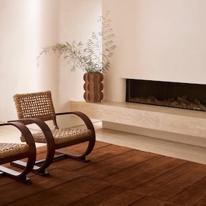May include: Two armchairs with dark brown curved wooden frames and woven tan seats and backrests, a brown textured vase containing green branches, and a large rectangular dark brown rug with visible horizontal seams on the floor.