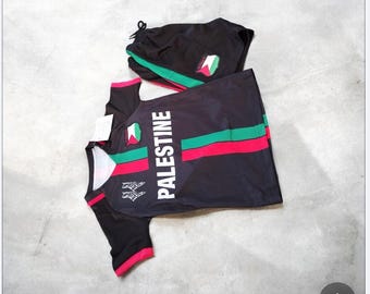 May include: Black athletic set comprising a short-sleeved shirt and shorts. The shirt displays the word "PALESTINE" in white, a red and green stripe, and a Palestinian flag emblem. The shorts have a matching emblem and green stripe.