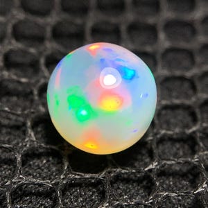 Könnte beinhalten: Ein runder, durchscheinender weißer Opal-Edelstein mit einem lebendigen Farbspiel, das deutliche Blitze in Neongrün, leuchtendem Orange und elektrischem Blau auf der glatten, polierten Oberfläche zeigt und das Licht mit einem weißen Glanzpunkt reflektiert.