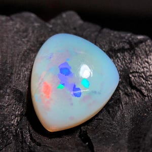 Könnte beinhalten: Ein dreieckiger Opal-Edelstein mit einer glatten, polierten Oberfläche. Der Stein hat eine durchscheinende, milchig-weiße Grundfarbe mit lebhaften Blitzen aus irisierendem blauem, grünem und orangefarbenem Licht auf der Oberseite.