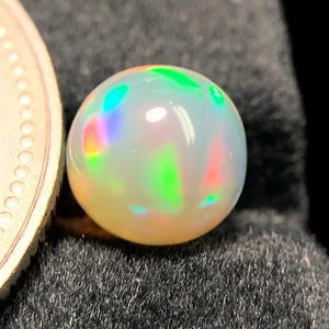 Könnte beinhalten: Ein runder, im Cabochon-Schliff gehaltener weißer Opal mit einem Farbspiel aus leuchtendem Grün, Orange und Pink. Der Edelstein ist neben einer Metallmünze platziert, um die Größe zu verdeutlichen, und zeigt eine glatte, polierte Oberfläche.