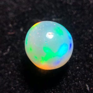 Könnte beinhalten: Ein runder, schillernder Opal-Edelstein. Der Opal zeigt ein Farbspiel mit Grün-, Blau- und Orangetönen. Der Edelstein ist vor einem dunklen Hintergrund platziert, der seine leuchtenden Farben und seine kugelförmige Form hervorhebt.