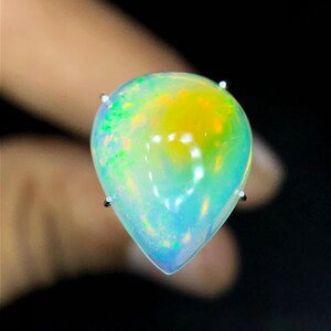 Könnte beinhalten: Ein birnenförmiger Opal-Edelstein mit einem durchscheinenden Körper und lebhaften Blitzen in Neongrün, Gelb und Orange. Der Stein ist mit kleinen silberfarbenen Krappen gefasst und weist eine glatte, polierte Oberfläche sowie einen Cabochon-Schliff auf.