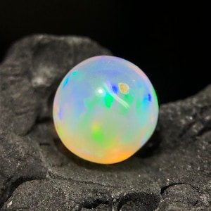 Könnte beinhalten: Ein runder, schillernder Opal-Edelstein mit milchig-weißer Grundfarbe. Der Opal zeigt Farbblitze in Blau, Grün und Gelb. Der Edelstein ist vor einem dunklen, strukturierten Hintergrund platziert.