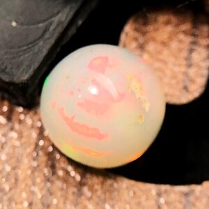 Könnte beinhalten: Ein runder, durchscheinender weißer Opal-Edelstein mit internen Farbspielen in Pink, Orange und Gelb. Der Stein hat eine glatte, polierte Oberfläche, die das Licht reflektiert und einen hellen Glanzpunkt auf dem oberen runden Bereich erzeugt.