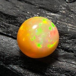 Könnte beinhalten: Ein runder, polierter Opal-Edelstein mit einem lebendigen Farbspiel. Der Opal hat eine warme orangefarbene Basis mit grünen und roten Lichtreflexen. Der Edelstein liegt vor einem dunklen, strukturierten Hintergrund.