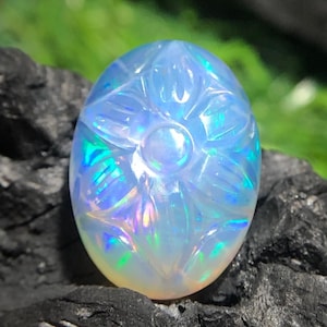 Könnte beinhalten: Ein ovaler, schillernder Opal-Edelstein mit einem geschnitzten Blumenmuster. Der Opal zeigt eine Farbpalette, darunter Blau, Grün und Gelb, vor einem dunklen, strukturierten Hintergrund. Der Edelstein ist der Fokus, der komplizierte Details zeigt.