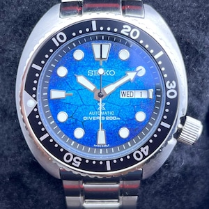 Puede incluir: Reloj de buceo automático Seiko de acero inoxidable con esfera azul. El reloj presenta un bisel giratorio negro, marcadores luminosos y una pantalla de fecha. La correa del reloj es de acero inoxidable.
