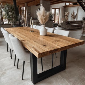 May include: A large, rectangular wooden dining table with a natural finish, supported by black metal legs. Six upholstered chairs with light gray fabric surround the table. A white vase with dried flowers sits on the table.