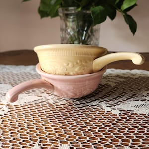 May include: Two small, ceramic bowls with handles, one stacked on top of the other. The top bowl is a pale yellow, and the bottom bowl is pink. Both bowls have a floral design and are sitting on a white lace tablecloth.