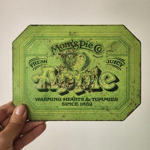 May include: A vintage green metal sign for Mom's Pie Co. with the word "Apple" in large, stylized letters. The sign also includes the phrases "Fresh," "Juicy," and "Warming Hearts & Tummies Since 1862."