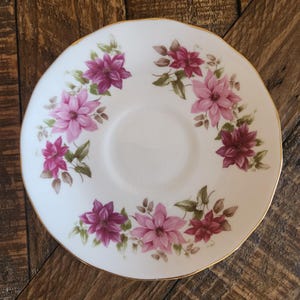 May include: White saucer with a gold rim, adorned with a floral pattern of pink and magenta flowers and green leaves. The flowers are arranged in a circular pattern around the center of the saucer. A vintage-style piece.