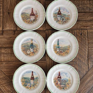 May include: Six white ceramic plates with a green rim. Each plate features a detailed illustration of a wine bottle with a vineyard scene. The plates are arranged on a wooden surface.